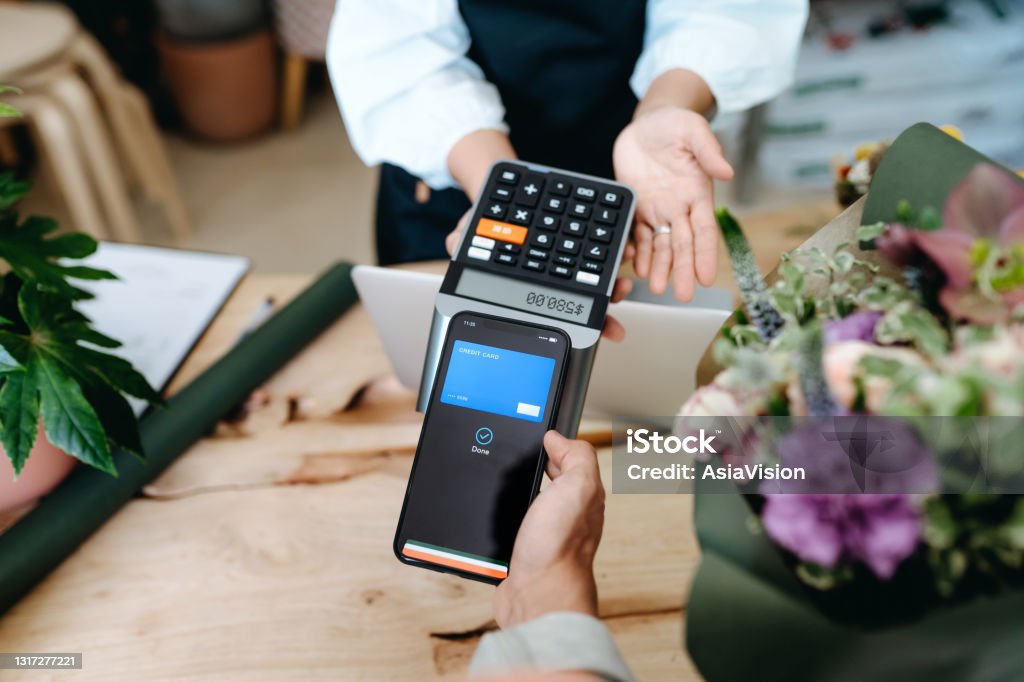 Personal perspective of young Asian man shopping at the flower shop. He is paying for a bouquet with his smartphone, scan and pay a bill on a card machine making a quick and easy contactless payment. NFC technology, tap and go concept Paying Stock Photo