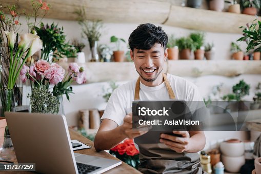 istock Asian male florist, owner of small business flower shop, using digital tablet while working on laptop against flowers and plants. Checking stocks, taking customer orders, selling products online. Daily routine of running a small business with technology 1317277259