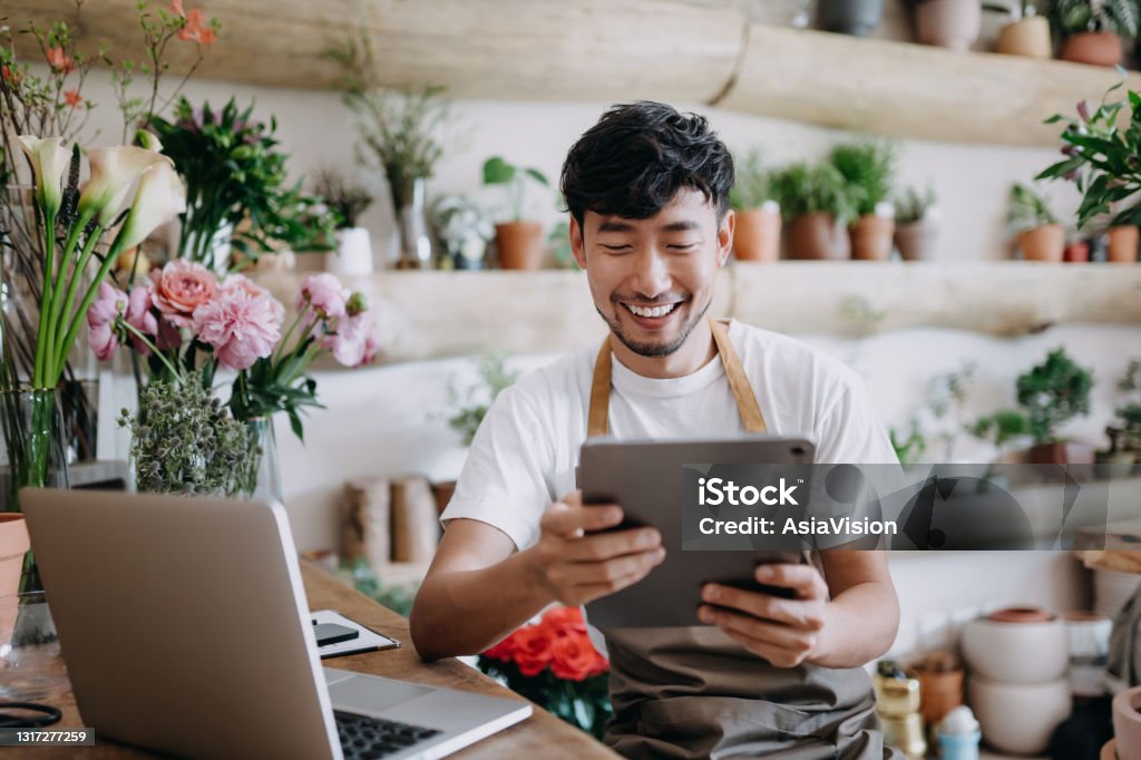 Asian male florist, owner of small business flower shop, using digital tablet while working on laptop against flowers and plants. Checking stocks, taking customer orders, selling products online. Daily routine of running a small business with technology Small Business Stock Photo