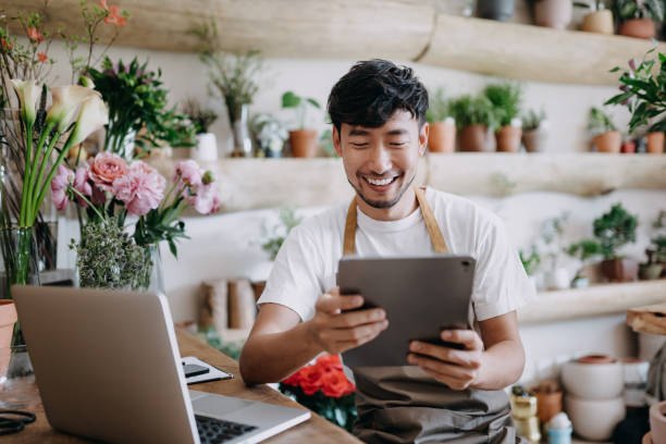 Asian male florist, owner of small business flower shop, using digital tablet while working on laptop against flowers and plants. Checking stocks, taking customer orders, selling products online. Daily routine of running a small business with technology stock photo