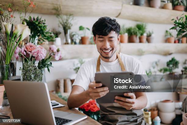 Asian Male Florist Owner Of Small Business Flower Shop Using Digital Tablet While Working On Laptop Against Flowers And Plants Checking Stocks Taking Customer Orders Selling Products Online Daily Routine Of Running A Small Business With Technology Stock Photo - Download Image Now