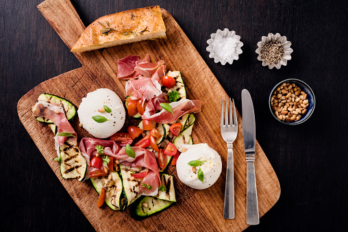 Overhead view of a salad lunch with; burrata, slices of grilled zucchini , parma ham, tomatoes, and toasted pine nuts. Burrata cheese is recognisable  by its loose texture: small, soft cheese curds and cream are packaged inside an outer shell made of mozzarella. Here served with warm alive oil and seasoned with salt and pepper. Colour, horizontal format with some copy space.