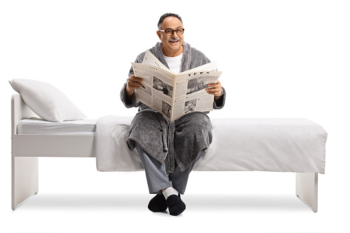 Mature man in  robe sitting on a bed and reading a newspaper isolated on white background