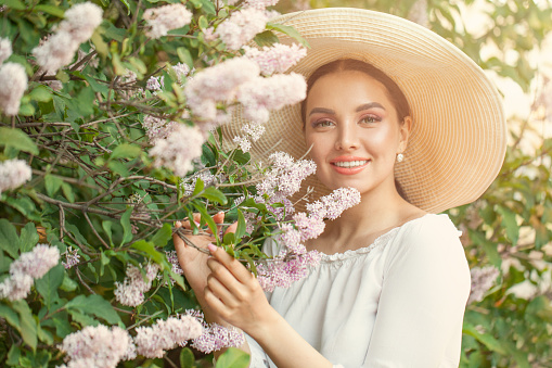 Portrait of cute young happy woman with spring flowers outdoors. Brunette female model in straw hat in flowers garden