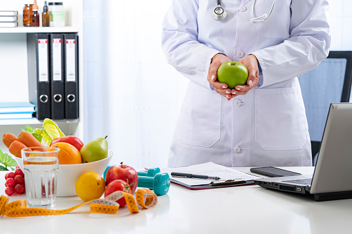 Right nutrition and diet concept. Nutritionist standing at office desk holding a green apple explaining the benefits of eating fruits and vegetables. An orange juice bottle, glass of water, tape measure are on the doctor's desk and complete the composition. Copy space available for text and/or logo. High resolution 42Mp studio digital capture taken with Sony A7rII and Sony FE 90mm f2.8 macro G OSS lens