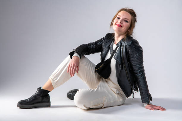 istock Cute woman in black leather jacket and white pants siting on the studio floor. 1317307199