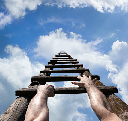 Muscular male hands gripping wooden ladder and reaching to the top, to the blue sky with white clouds and light, success concept