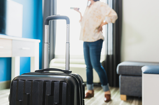 A photo of a suitcase packed and ready in the foreground, with the unrecognizable woman in the background using her smart phone for a quick call before leaving.