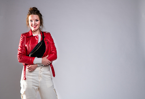 Young woman wearing travel fanny pack, white pants and red leather jacket, standing, posing against grey background. Studio shoot. She is smiling and looking at the camera.