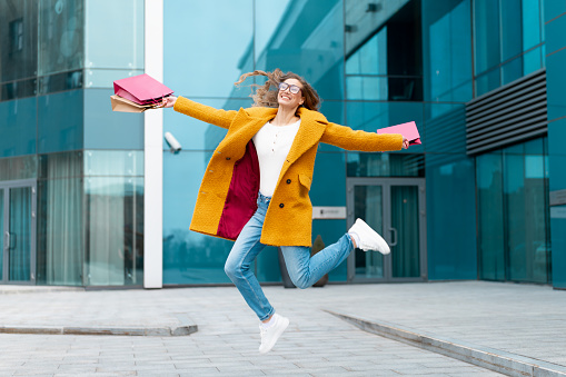 Business Woman With Shopping Bags Dressed Yellow Coat Jumping With Happiness Outdoors Corporative Building Caucasian Female Business Person Near Office Building Stylish Businesswoman Season Sale