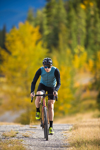 A young man goes for a gravel road bicycle ride in autumn. Gravel bicycles are similar to cyclo-cross bikes with sturdy wheels and tires for riding on rough terrain. He is wearing a cycling helmet, sunglasses and road cycling clothing.