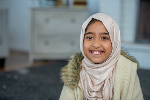 A girl wearing a hijab smiles at the camera as she sits in her living room.