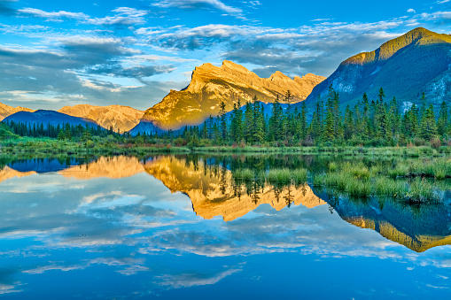 A scenic view of a lake reflecting rocky mountains covered with green pine trees on a sunny day