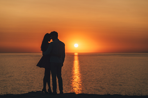 The young couple standing on the sea shore on the picturesque sunset background