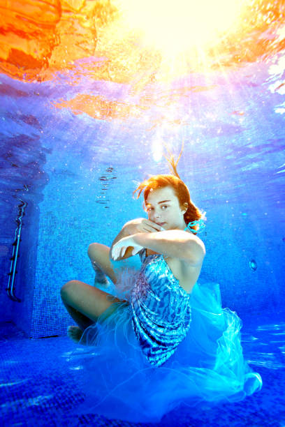 retrato de uma jovem brincalhão que nada e posa para a câmera sob a água na piscina em um vestido azul brilhante, nos raios brilhantes do sol. conceito. orientação vertical. - teenage girls ideas blue yellow - fotografias e filmes do acervo