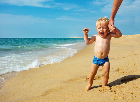 Little toddler boy walk a bit afraid near sea waves on the beach holding mother by hand