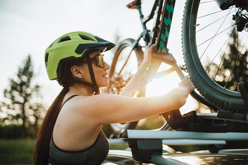 A side view of a young woman with helmet attaching her mountain bike on a roof carrier on a car.