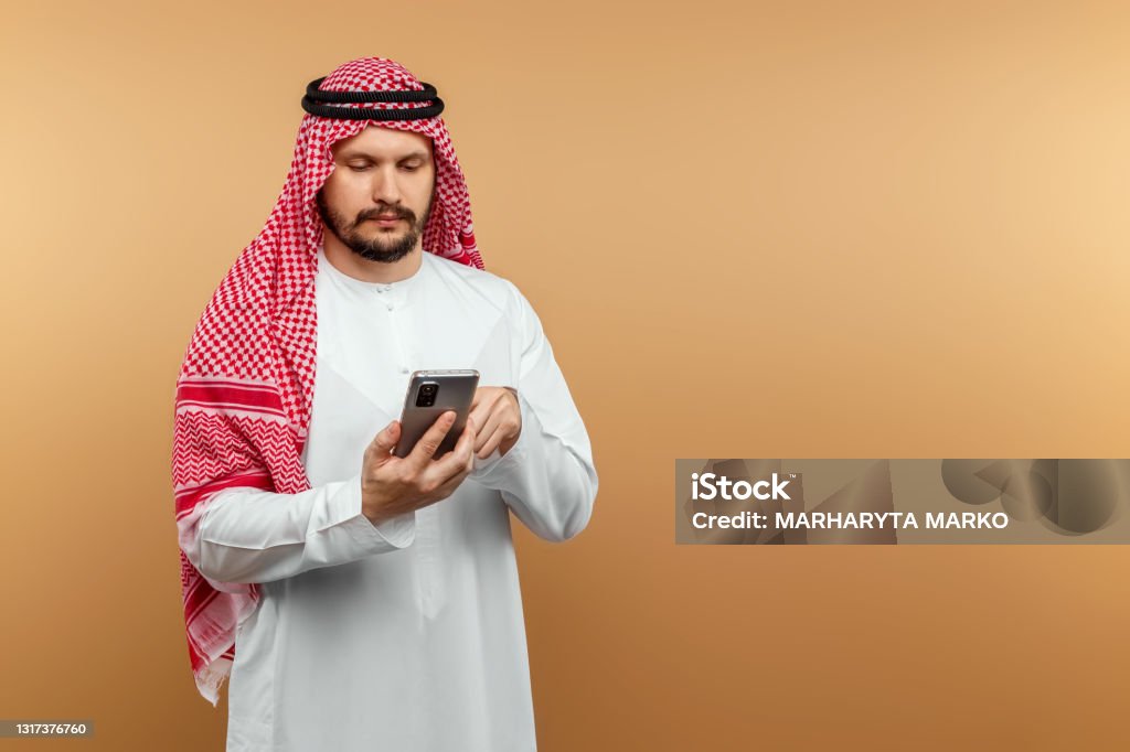 Man businessman in national dress holds a smartphone in his hands, beige background. Dishdasha, kandora, thobe, middle east traditional menswear concept, . Copy space. Man businessman in national dress holds a smartphone in his hands, beige background. Dishdasha, kandora, thobe, middle east traditional menswear concept, . Copy space Men Stock Photo