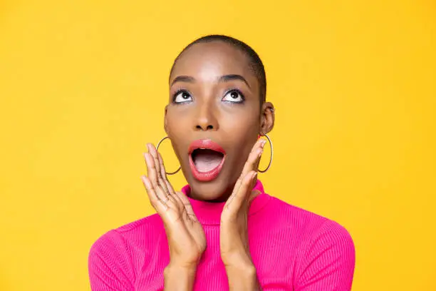 Close up portrait of surprised young African American woman looking upward with hands cupped around mouth isolated on studio yellow background