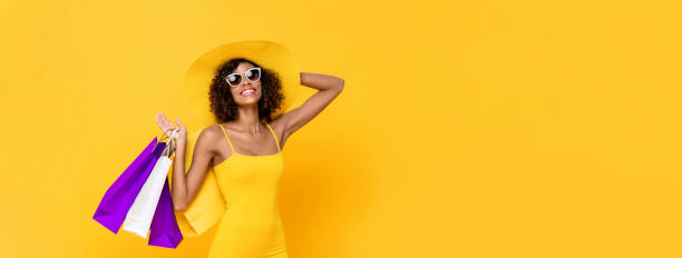 복사 공간이있는 고립 된 스튜디오 노란색 배너 배경에서 모자와 쇼핑 백을 들고 선글라스를 착용한 쾌활한 미소 짓는 아프리카 계 미국인 여성의 여름 초상화 - sunglasses women studio shot isolated 뉴스 사진 이미지