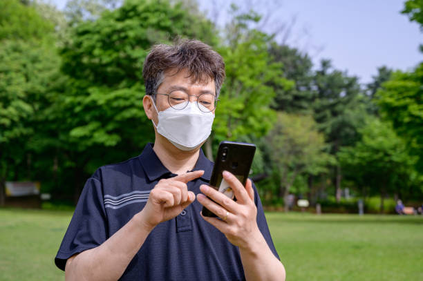 a middle-aged asian man wearing a face mask and using a smartphone on the lawn. - 2127 imagens e fotografias de stock