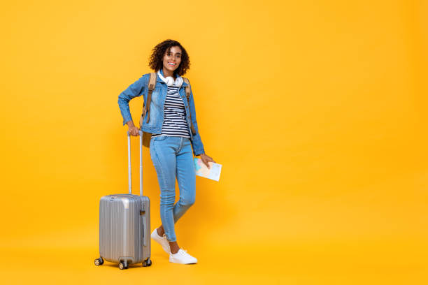 黄色のスタジオの背景に立って荷物を保持している笑顔の若いアフリカ系アメリカ人女性バックパッカーの完全な長さの旅行の肖像画 - female mobility blank teenage girls ストックフォトと画像