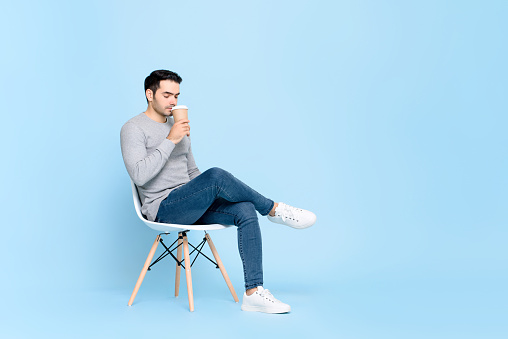 Portrait of young handsome caucasian man taking a break sitting calmly sipping coffee in isolated studio blue background