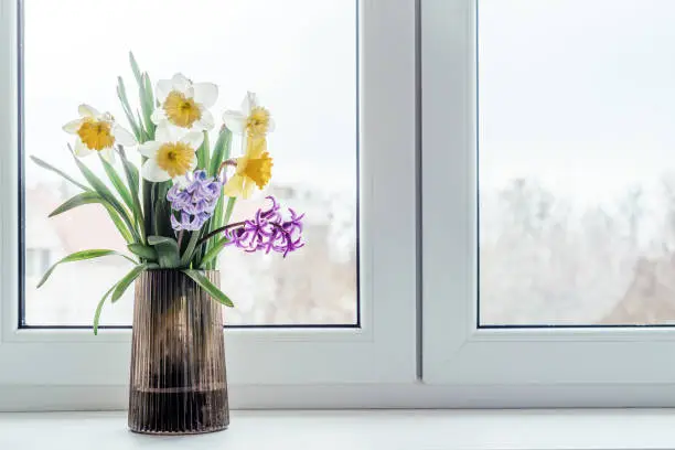 Photo of Spring bouquet of yellow narcissus and blue and purple hyacinth in glass vase on window sill