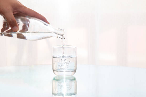 ボトルからガラスに新鮮なろ過精製水を注ぐ男の手を閉じると、飲み物のためのテーブルに反射します - purified water bottle water drink ストックフォトと画像