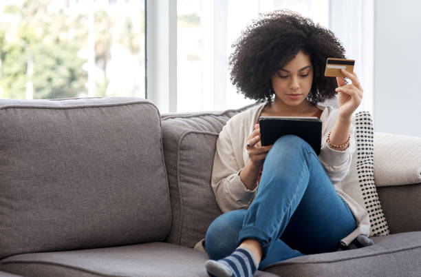 shot of a young woman shopping online while sitting on the couch at home - money to burn fotos imagens e fotografias de stock