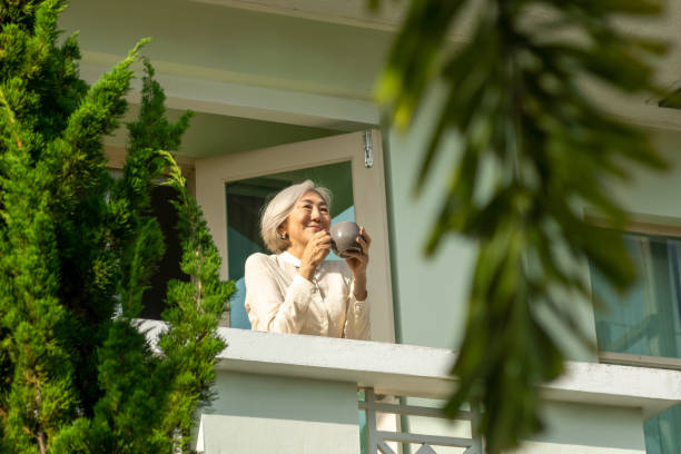 Mature asian woman standing on balcony with cup of coffee. Morning, lifestyle concept, text space stock photo