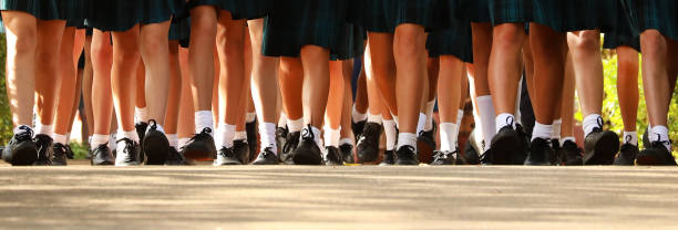 wide image of a large amount of female students walking together into school. lower leg only wide worms eye view multiple many female girls legs wearing school uniform of white socks black shoes and tartan skirt. Walking in together on the first day of high school. school uniform stock pictures, royalty-free photos & images