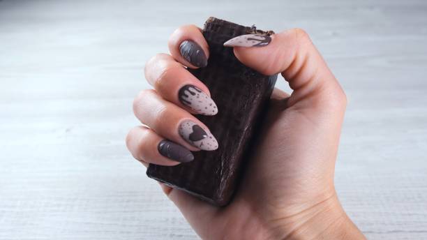 mains femelles avec de beaux ongles bruns de manucure sous forme de bonbons de chocolat, retenant une barre dépliée de chocolat noir, plan rapproché. profitez du dessert - brown chocolate candy bar close up photos et images de collection