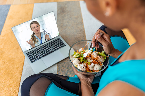 Over the shoulder shot of a patient talking to a doctor using of a mobile phone, woman eating salad from a bowl while sitting on the floor.