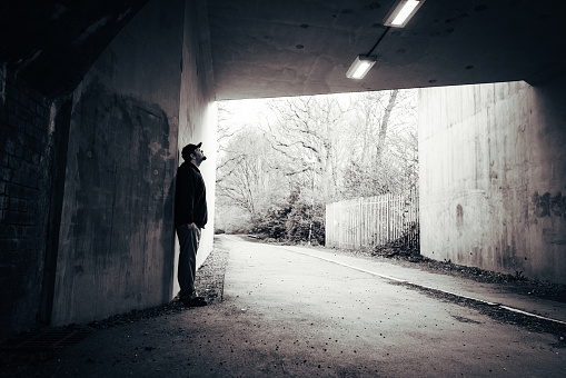 Monochrome portrait of a mid adult lonely man standing alone in a tunnel in the suburbs of a town. He is standing, looking forlorn, with his back against the concrete wall.