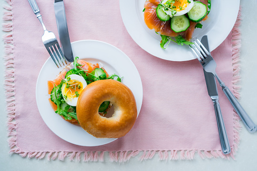 Top view bagel sandwiches with salmon, soft egg, salad, microgreen sprouts, cucumber and cream cheese served on pink napkin with cutlery on the white kitchen table. Healthy Breakfast. Selective focus