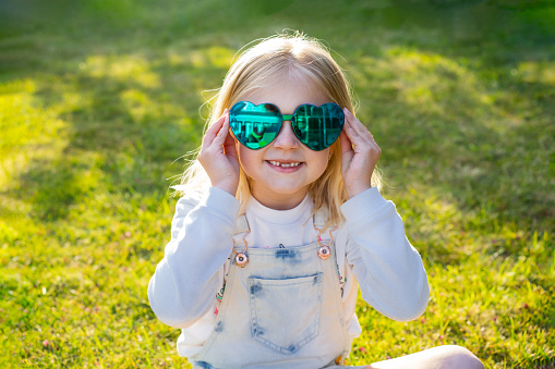 Outdoor portrait of a little blonde smiling toothless girl wearing heart-shaped sunglasses, sitting on fresh green grass. Party for children, summer fun, happy childhood