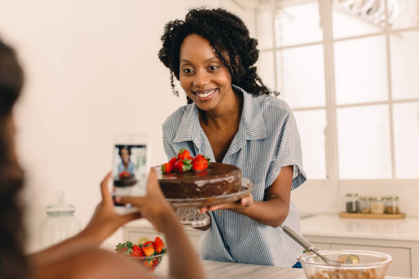 Woman with cake being photographed by daughter Woman holding a cake being photographed by her daughter at home. Girl taking photo with mobile phone of her mother with cake in kitchen. confectioner photos stock pictures, royalty-free photos & images