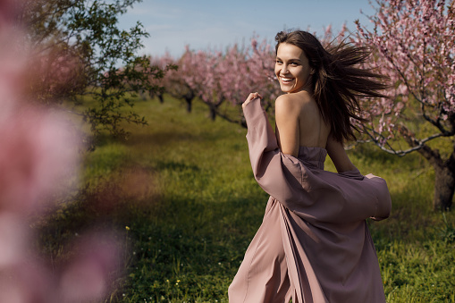 Beautiful brunette outdoors alone in a blooming pink peach garden. Beautiful girl on the street in a brown dress, spring portrait. Young woman in a peach garden, spring concept. Happy girl walking outdoors in the spring garden.