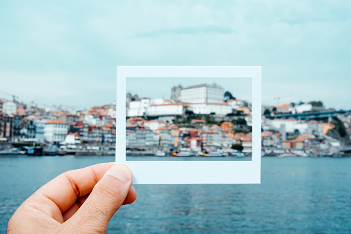 closeup of a young caucasian man with a cardboard white frame in his hand, simulating an instant photograph, at the Douro River framing the Episcopal Palace and the Ribeira district of Porto, Portugal
