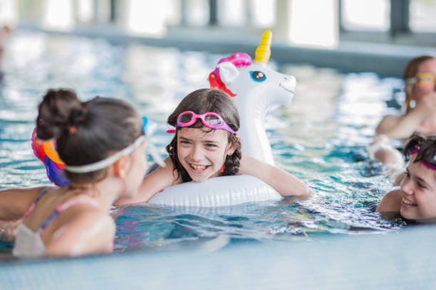 Happy girl floating in swimming pool on inflatable unicorn and talking to her friends stock photo
