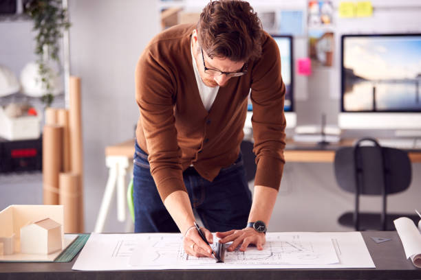 istock Male Architect Standing At Desk In Office Amending Plan Or Blueprint With Ruler 1317442811
