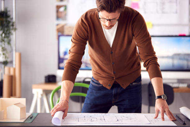 istock Male Architect In Office Unrolling Building Plan Or Blueprint On Desk 1317442864