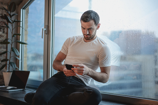 Handsome young brunette man in white t-shirt sitting on the windowsill at home on a rainy day with a phone in hands
