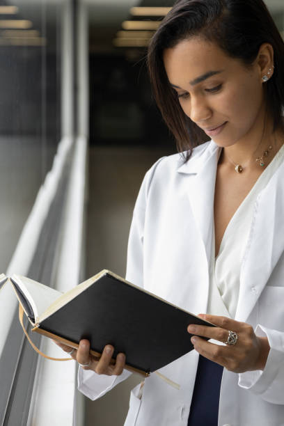 брюнетка молодая женщина-врач с короткими волосами, стоя читая книгу, носить белое пальто, профессиональный - book reading white women стоковые фото и изображения