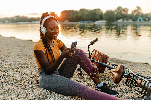 Young African American woman is outdoors, she is smiling and enjoying a nice summer day while holding a mobile phone. Her bicycle is next to her