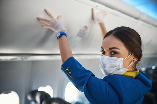 Airline stewardess in protection mask turning her head and looking into camera while pressing hands to overhead bin cover