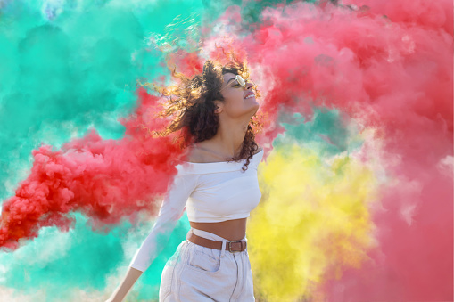 Beautiful young woman surrounded  light up colored smoke bombs - Happy friends having fun in the park with multicolored smoke bombs - Young students celebrating spring break together. Holi festival.