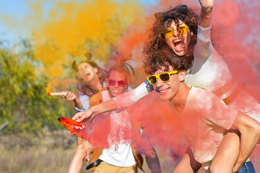 Beautiful young man and woman hold light up colored smoke bombs - Happy friends having fun in the park with multicolored smoke bombs - Young students celebrating spring break together. Holi festival.