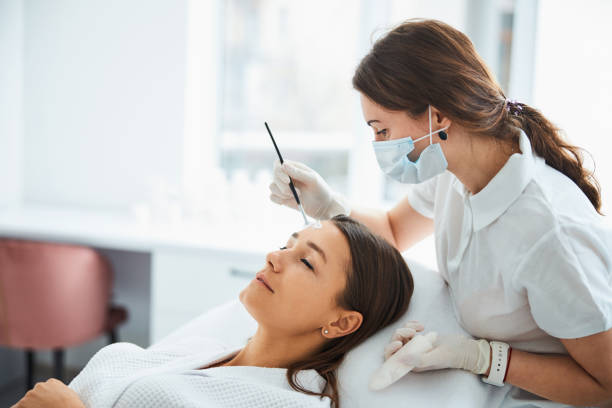 Dermatologist applying a face mask to the patient skin Concentrated skilled female beauty therapist spreading a cosmetic product over a young Caucasian woman forehead beautician stock pictures, royalty-free photos & images